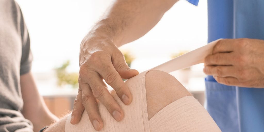 hands-of-clinician-in-blue-uniform-wrapping-injured-knee-of-young-patient.jpg